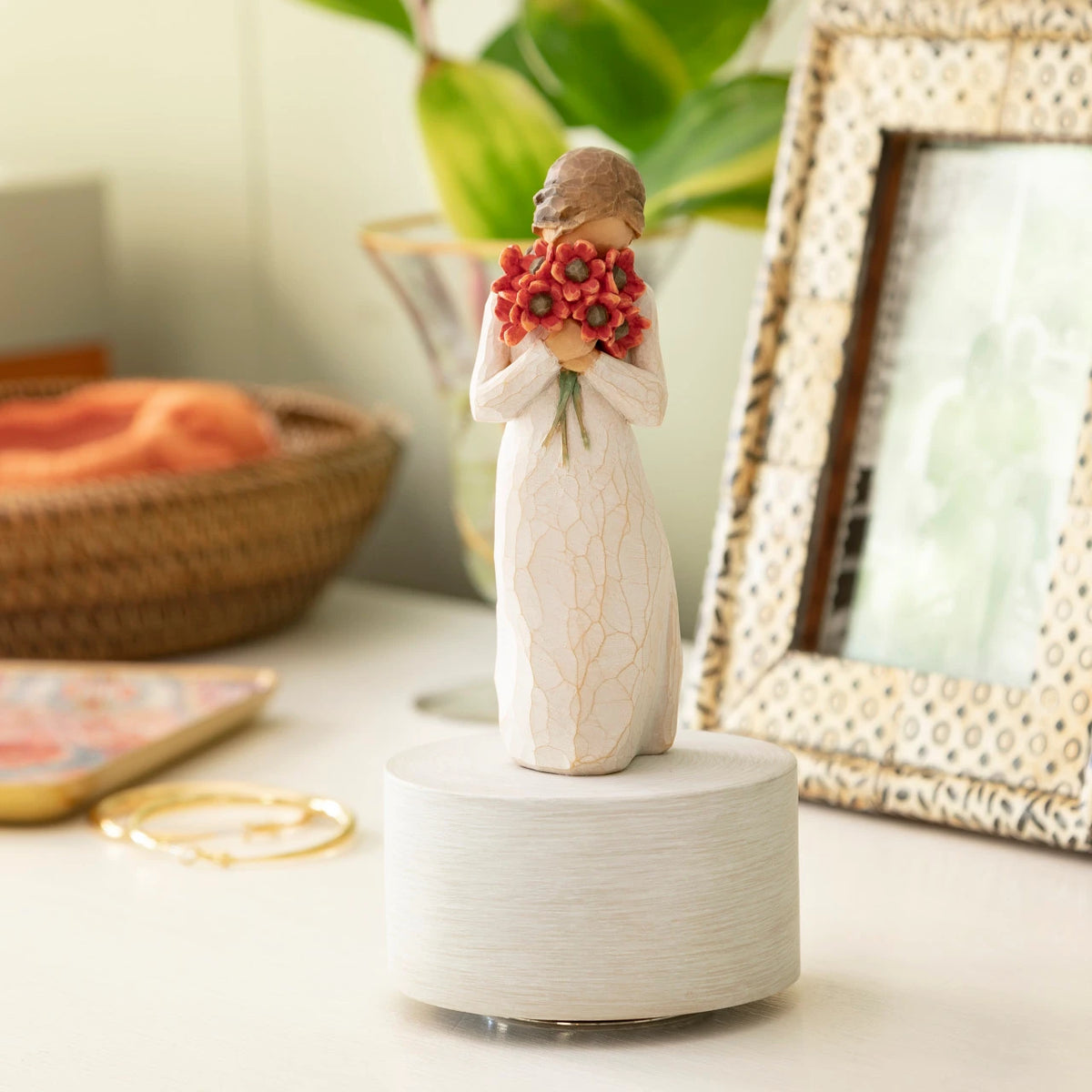 Willow Tree Surrounded by Love Musical, 7.5”h hand-painted resin figure in cream dress holding large bouquet of red-orange poppies up to face, atop round, cream base containing musical element, on desk with photos