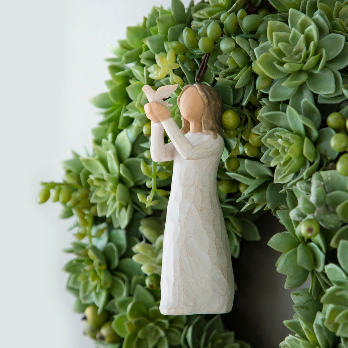 Willow Tree Soar Ornament, 4.5”h hand-painted resin figure in cream dress, holding white dove in extended arms, brown elastic cord, hanging in wreath of succulents