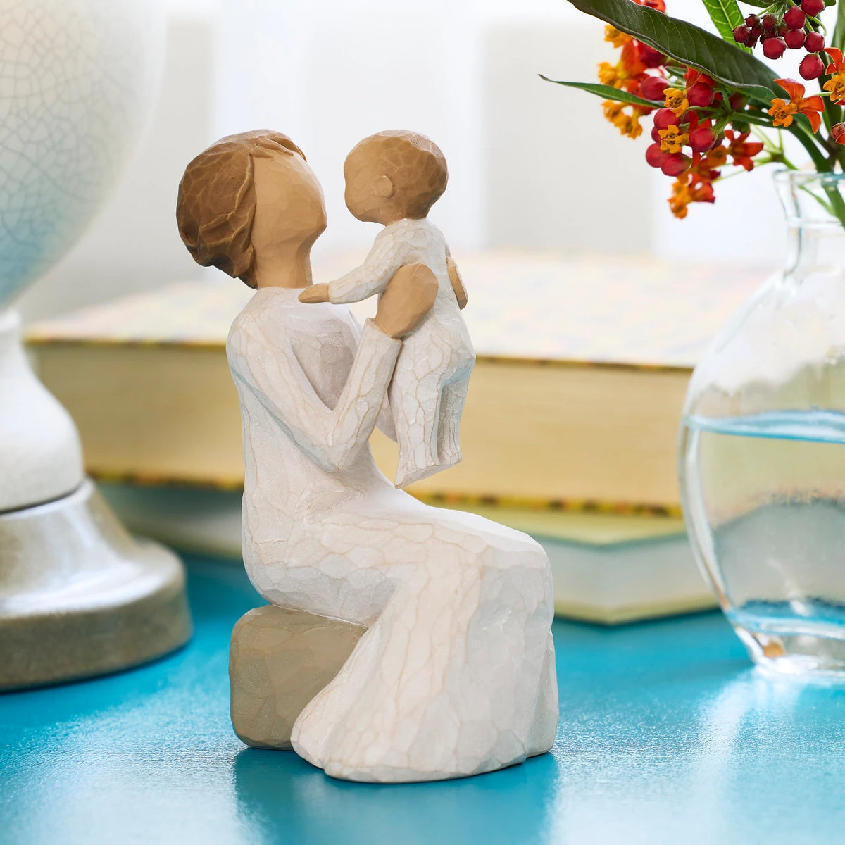 Willow Tree Grandmother, 5.5”h hand-painted resin figure of seated older woman in cream dress, holding baby up in the air, on blue shelf with books