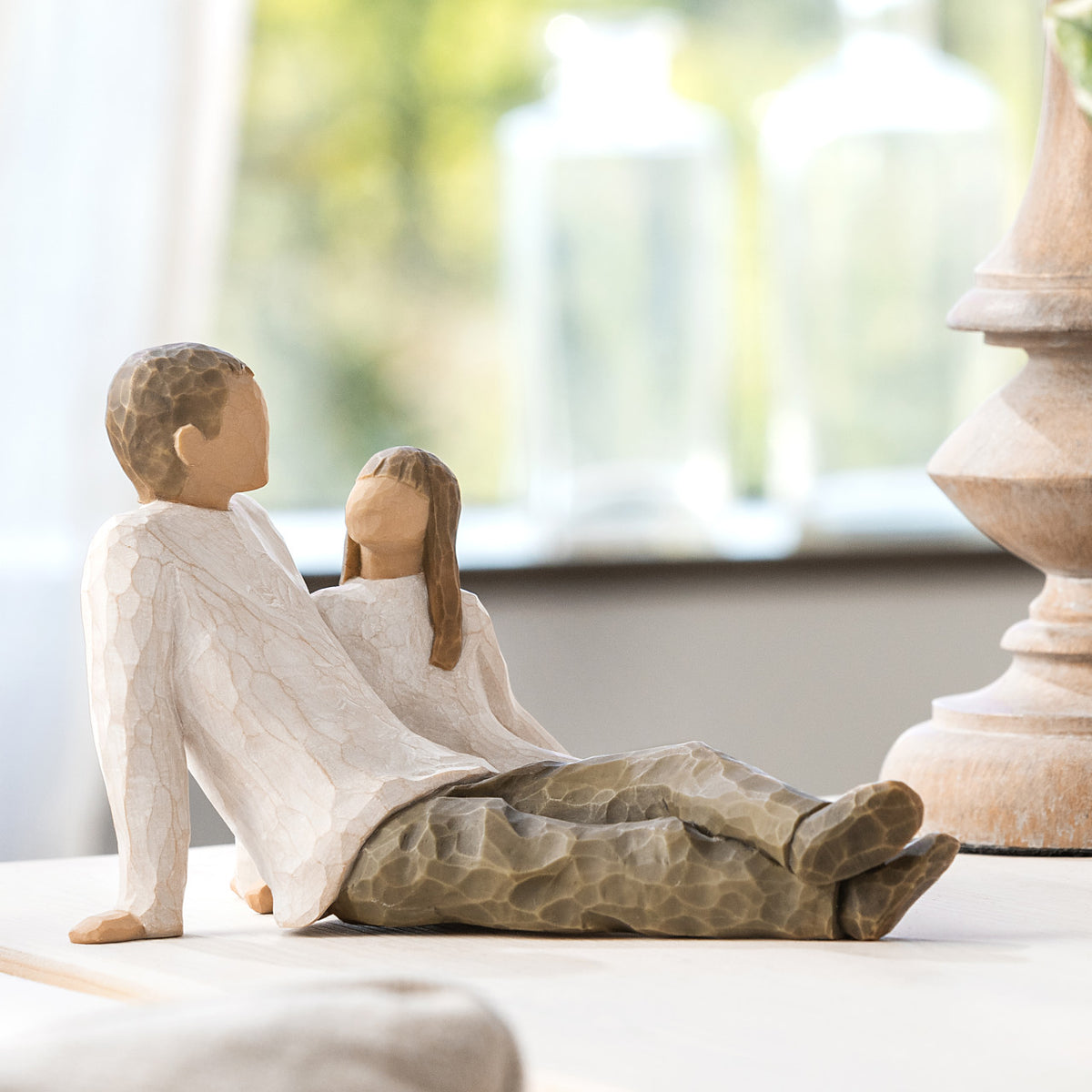 Willow Tree Father and Daughter, 5”h hand-painted resin figure of seated man dressed in neutral colors, legs outstretched, daughter in cream dress by his side, on table by window