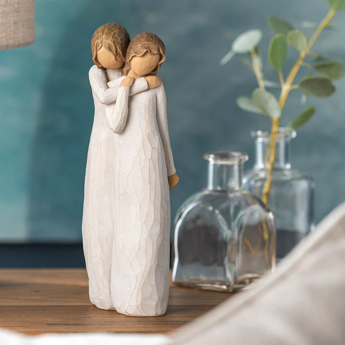 Willow Tree Chrysalis, 9”h hand-painted standing resin figure of two women in cream dresses; one standing behind and embracing the other, on table with glass vases