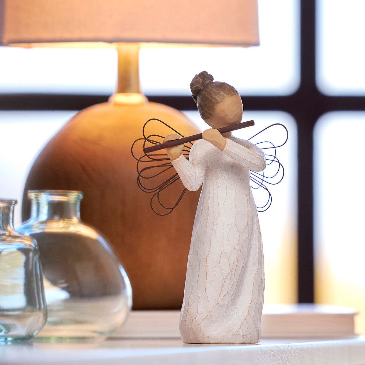 Willow Tree Angel of Harmony, 6"h hand-painted standing resin figure in cream dress with wire wings, playing the flute, on shelf with glass bottles and warm light