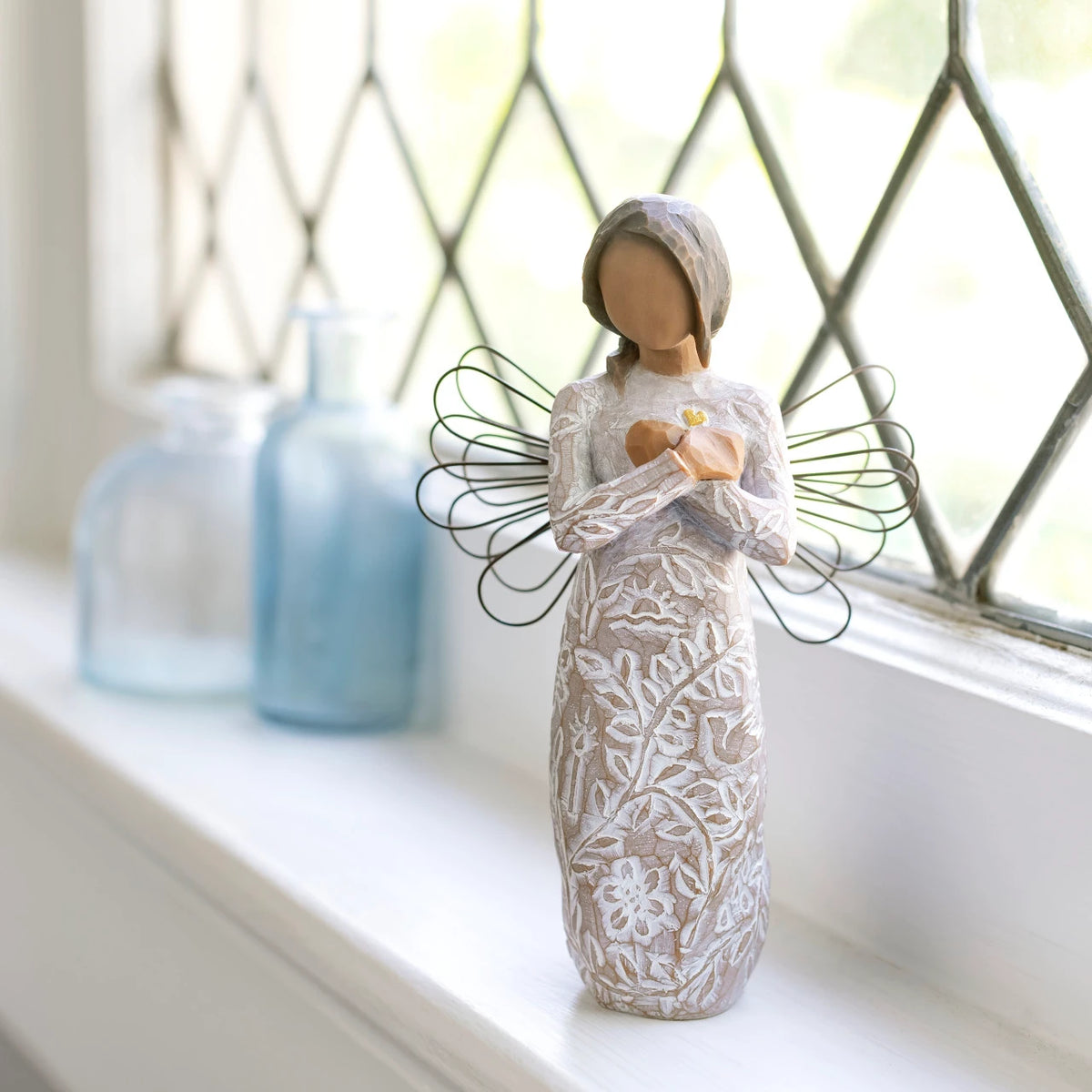 Willow Tree Remembrance (darker skin), 5.5”h hand-painted standing resin angel figure with wire wings, arms crossed over gold heart, in cream dress carved with nature icons, on bright windowsill