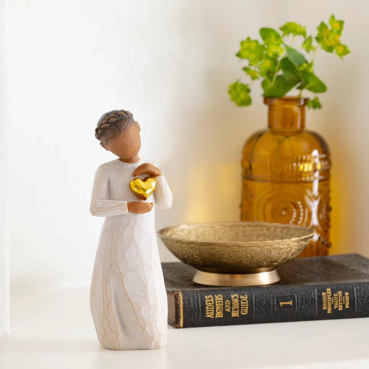Willow Tree Keepsake, 5.5”h hand-painted standing resin figure with darker skin and hair, in cream dress, framing gold heart with both hands on her chest, on desk with vase