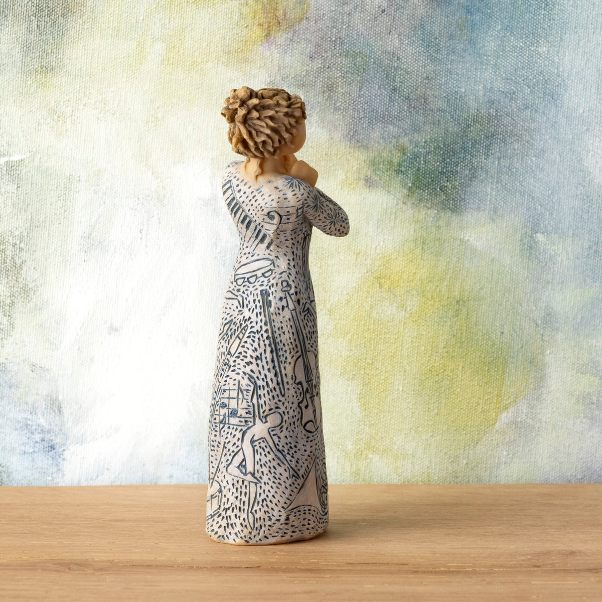 Willow Tree Music Speaks (lighter skin), back view of 7”h hand-painted standing resin figure wearing dress carved with symbols of music and dancing, in front of blue-green painting