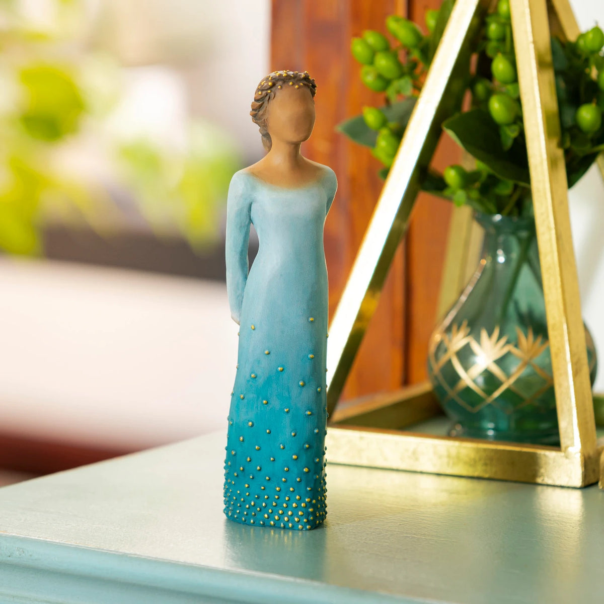 Willow Tree Radiance, 7.5”h hand-painted standing resin figure with darker skin and hair, in deep blue ombré dress dotted with gold leaf, arms clasped behind back, on table near window