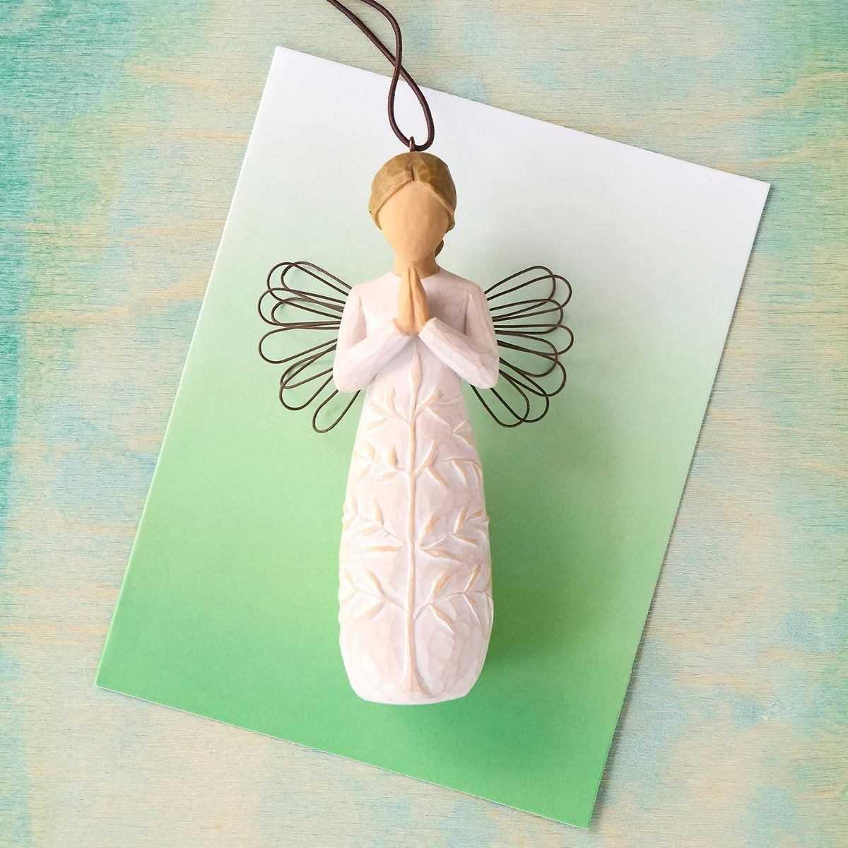 Willow Tree a tree, a prayer Ornament, 4”h hand-painted resin angel figure in cream dress with wire wings, hands in prayer position, tree carving on dress, brown elastic cord, laying in front of green painting