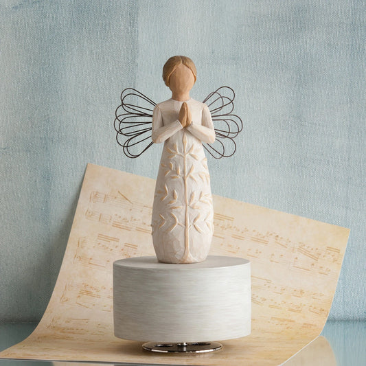 Angel figurine in white dress with hands folded in prayer - she has black wire wings and is standing on round white plaque - background is light blue with a tan colored music sheet below her