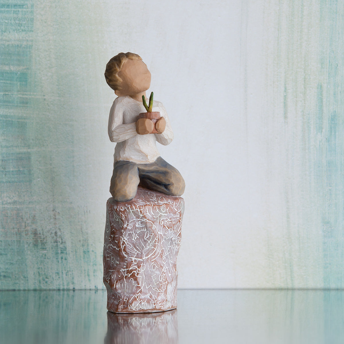 Willow Tree something special, 5.5”h hand-painted figure of small boy holding plant in pot, kneeling on tall rock etched with nature imagery, in front of grey painting