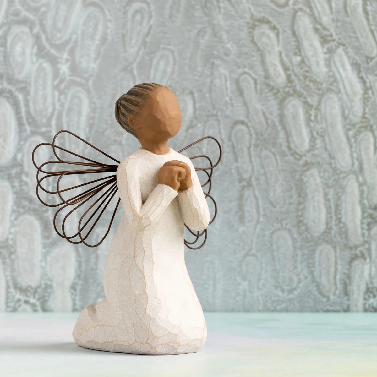Willow Tree Angel of the Spirit, 4"h hand-painted resin kneeling figure with darker skin and hair, cream dress with wire wings, looking upwards with hands in prayer position. Silver painting in background