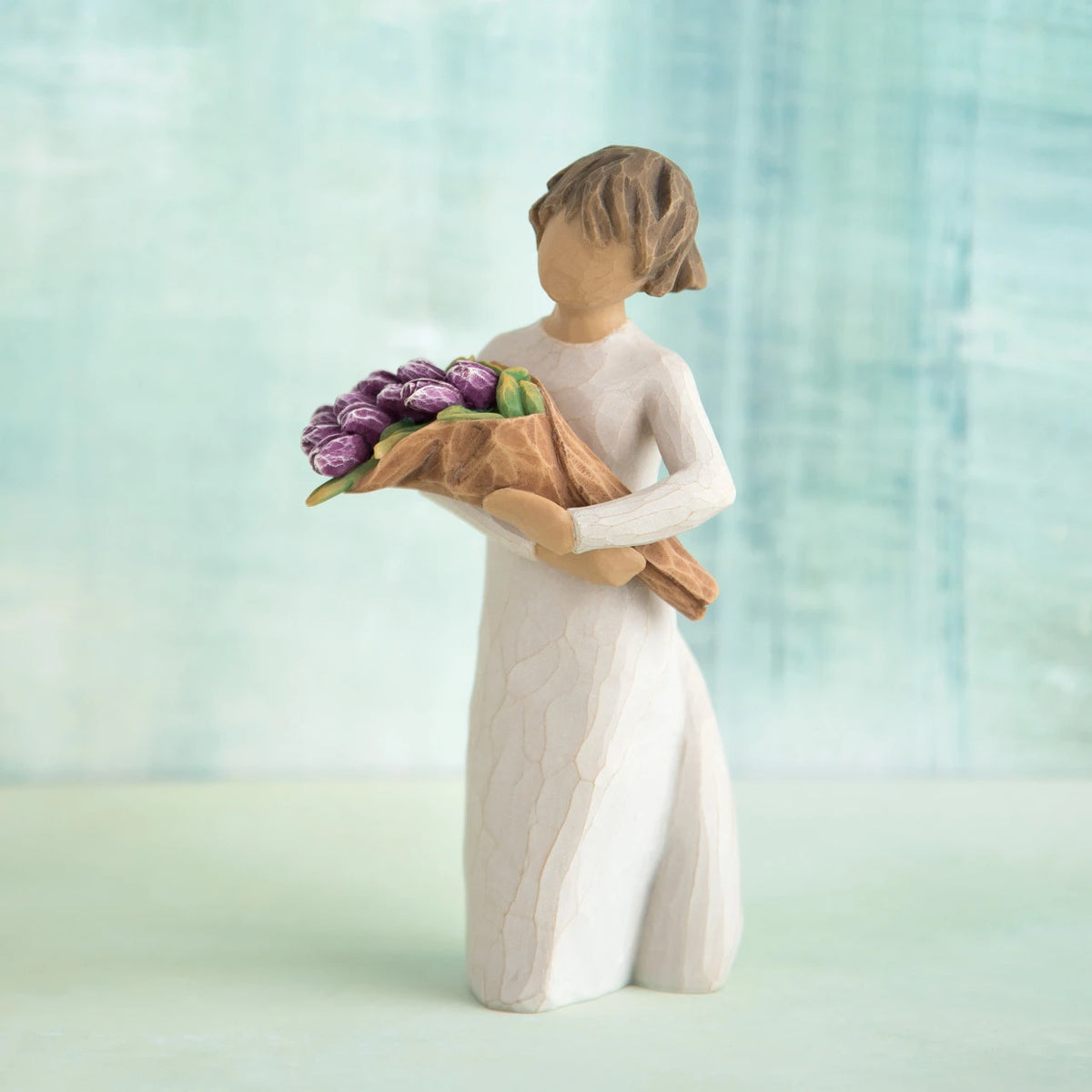 Willow Tree Surprise, 5”h hand-painted standing resin figure in cream dress, holding bouquet of purple tulips, in front of green-blue painting