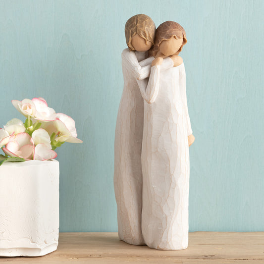 Willow Tree Chrysalis, 9”h hand-painted standing resin figure of two women in cream dresses; one standing behind and embracing the other, on wooden shelf with flowers
