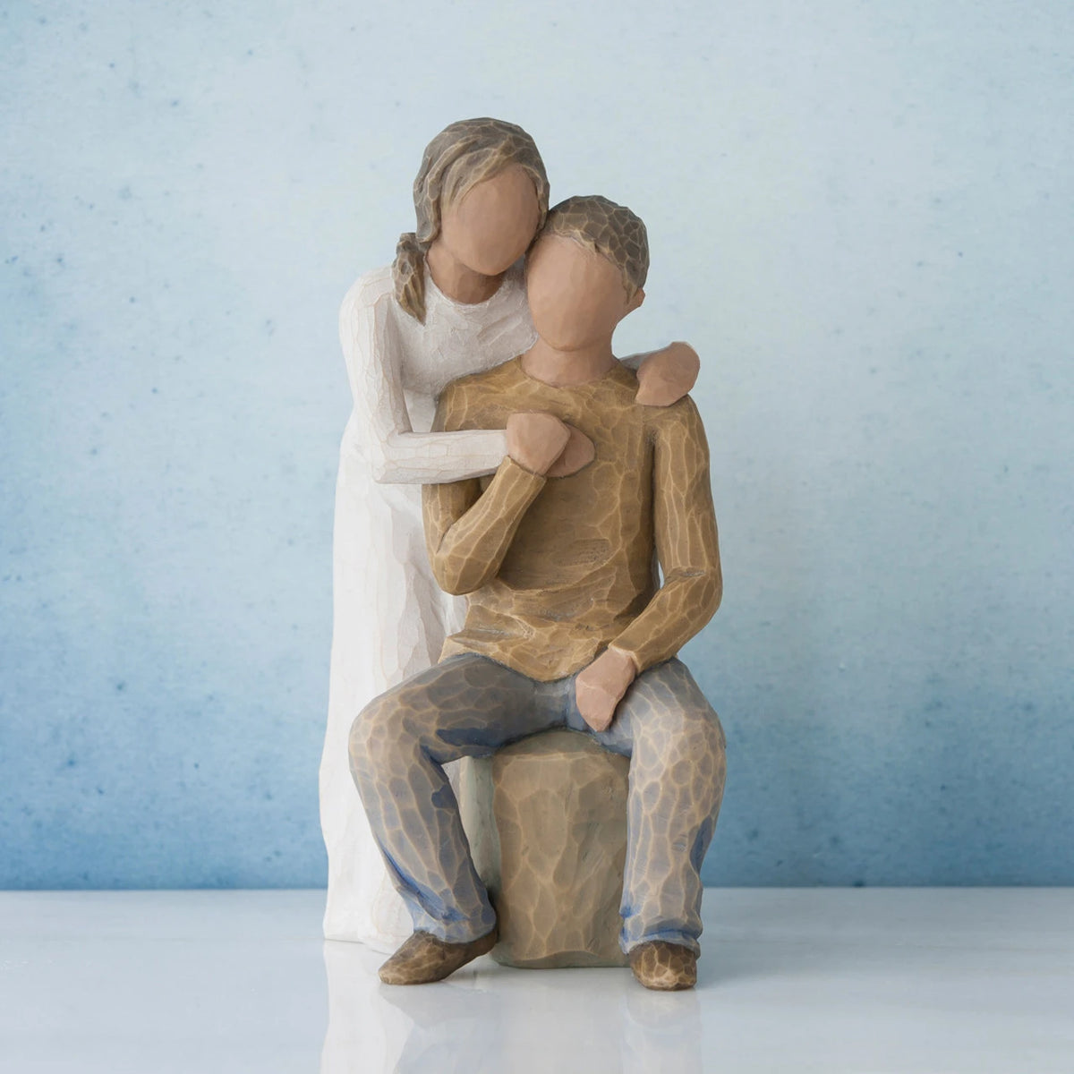 Man figurine sitting on rock wearing brown shirt and blue jeans with woman figurine in white dress with arms wrapped around him - blue background