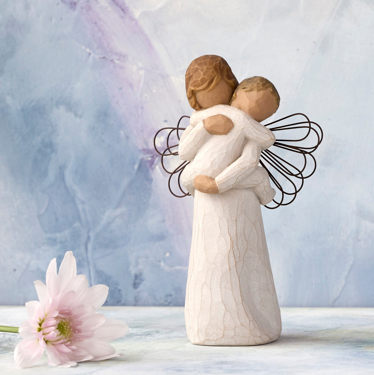 Willow Tree Angel's Embrace, 5"h hand-painted standing resin figure in cream dress with wire wings, holding baby in arms, in front of blue painting