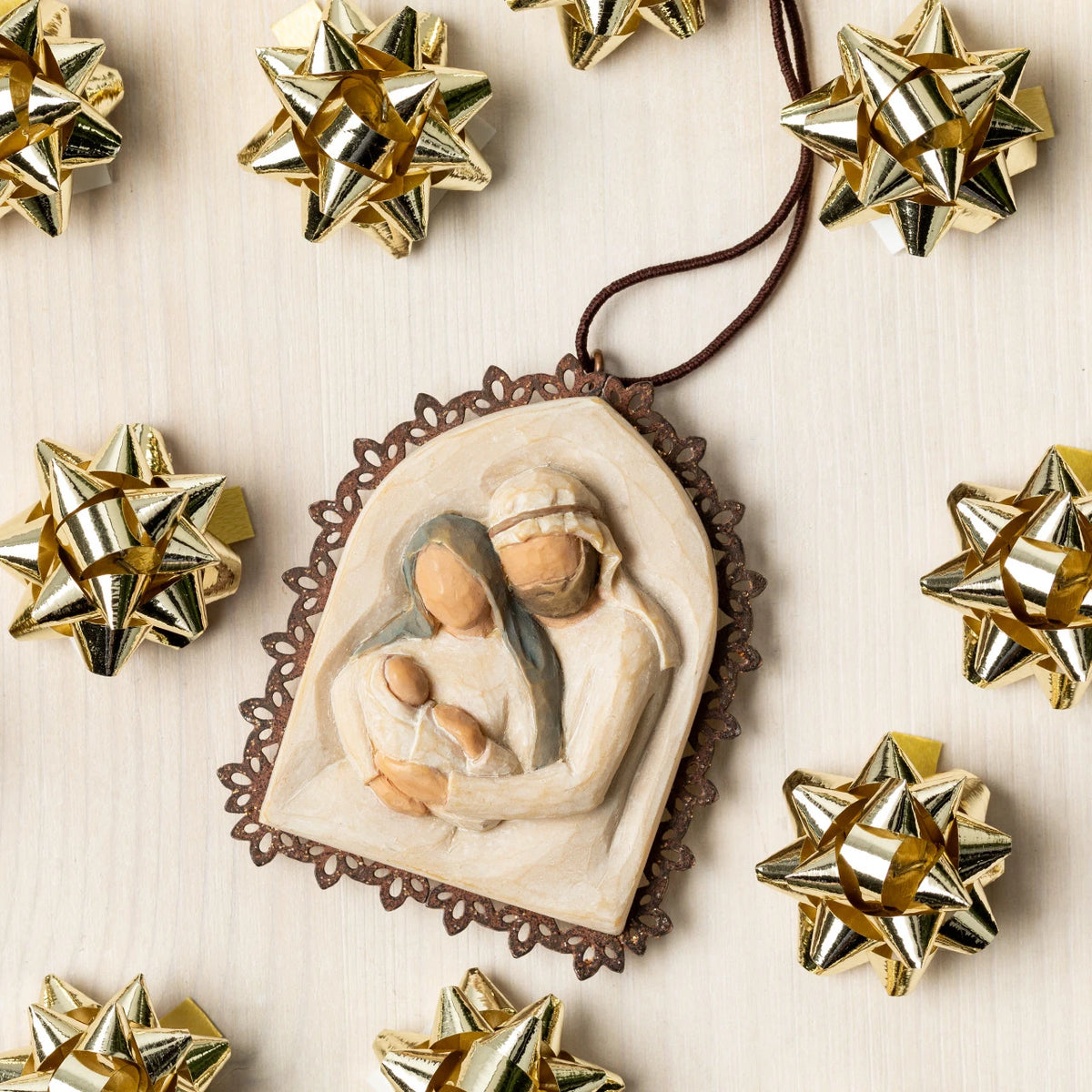 Willow Tree Holy Family Metal-edged Ornament, 2”w x 2.25”h hand-painted resin bas-relief carving of Joseph and Mary holding baby Jesus, edged in pierced-metal filigree, brown elastic cord for hanging, arranged with gold gift bows