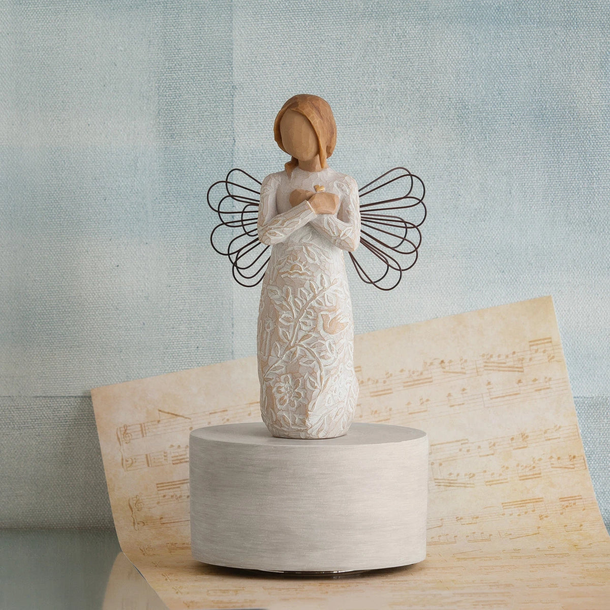 Standing faceless angel figurine in white dress with black wire wings - she is standing on round white stand with music sheet and blue background