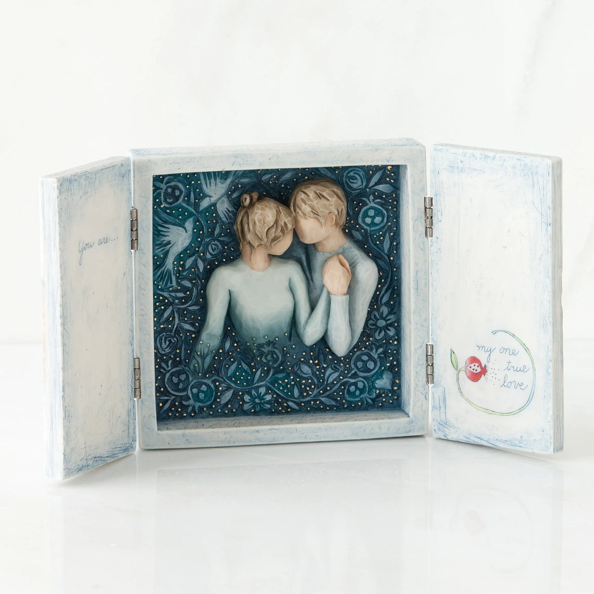 Open light wooden book with three parts - center is blue sky with two carved in faceless boy and girls hugging one another