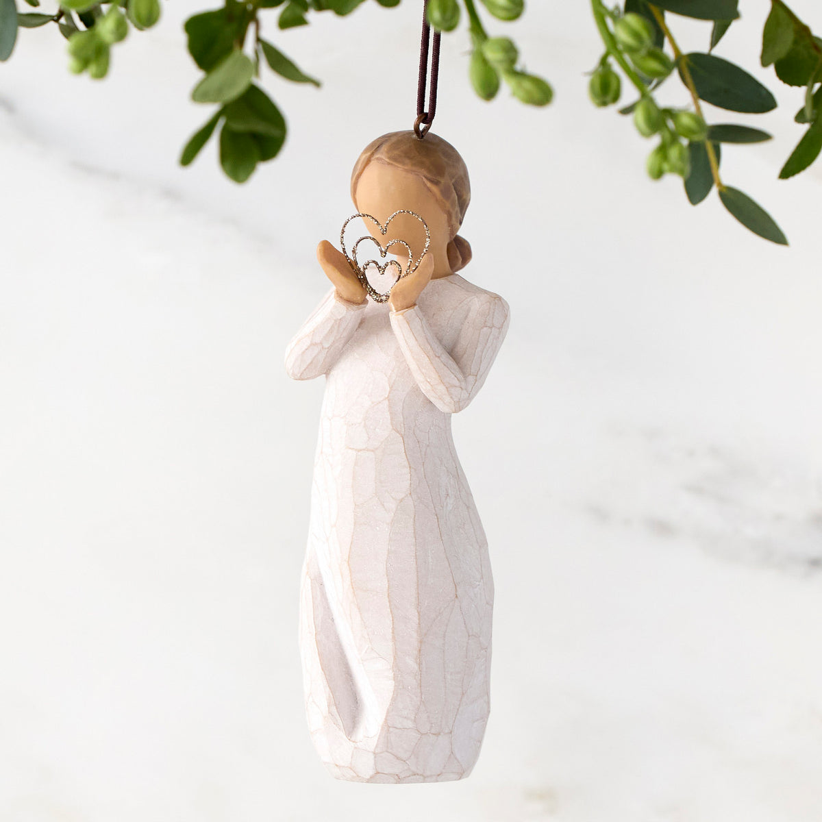 Figure in cream dress, holding thin wire concentric heart in hands. Wire heart is dusted with gold glitter. Hook and loop affixed to figure's head