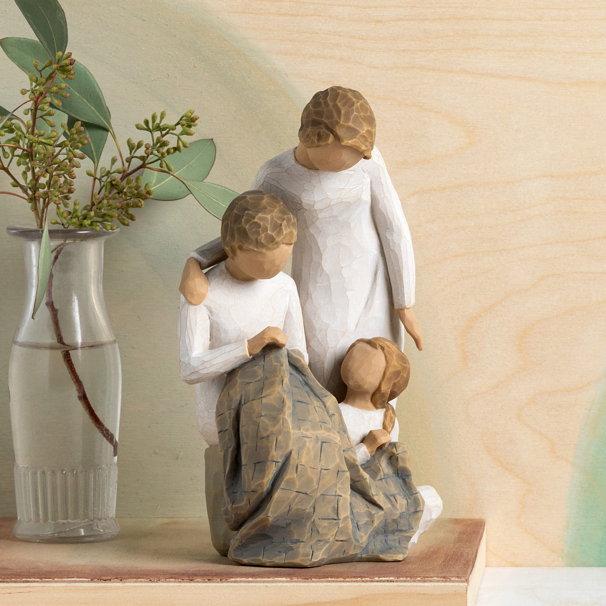 Willow Tree Generations, 7”h hand-painted resin figure of seated older woman sewing blue quilt on her lap; standing woman at her shoulder; seated young girl at her feet, all in neutral clothing, on table with green florals