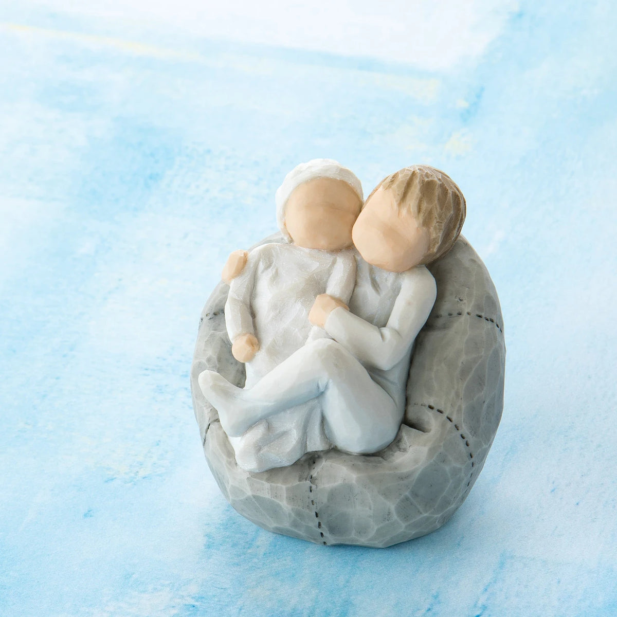 Willow Tree My new baby (sky), 2.5”h hand-painted resin figure of toddler in blue onesie holding infant in cream, cuddled together in a beanbag, in front of blue painting
