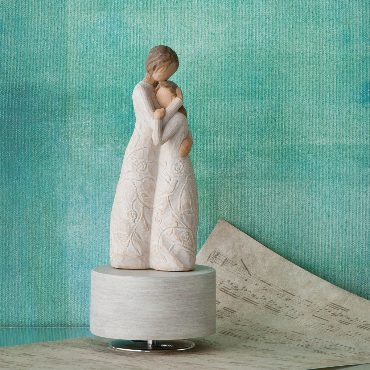 Willow Tree Close to Me Musical, 7.5”h hand-painted resin figure of woman embracing tween girl, both in cream dresses carved with intertwined vines and leaves, atop round, cream base containing musical element, in front of deep teal painting