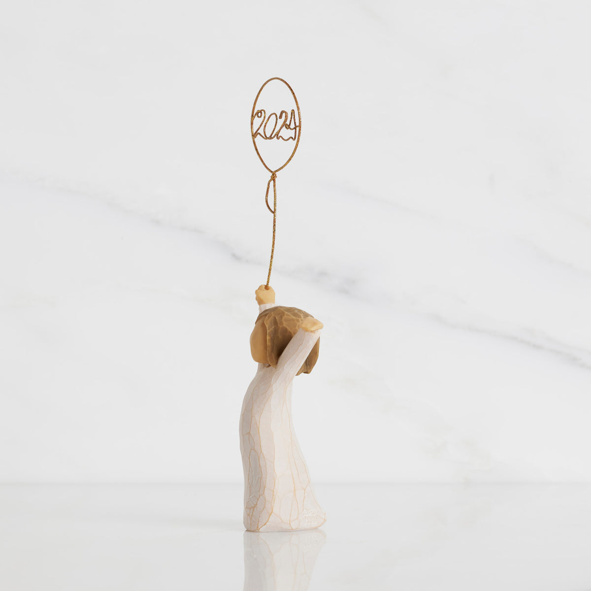 Facing left: standing figure in cream dress, arms raised, holding gold wire balloon with 2024 written in gold wire inside