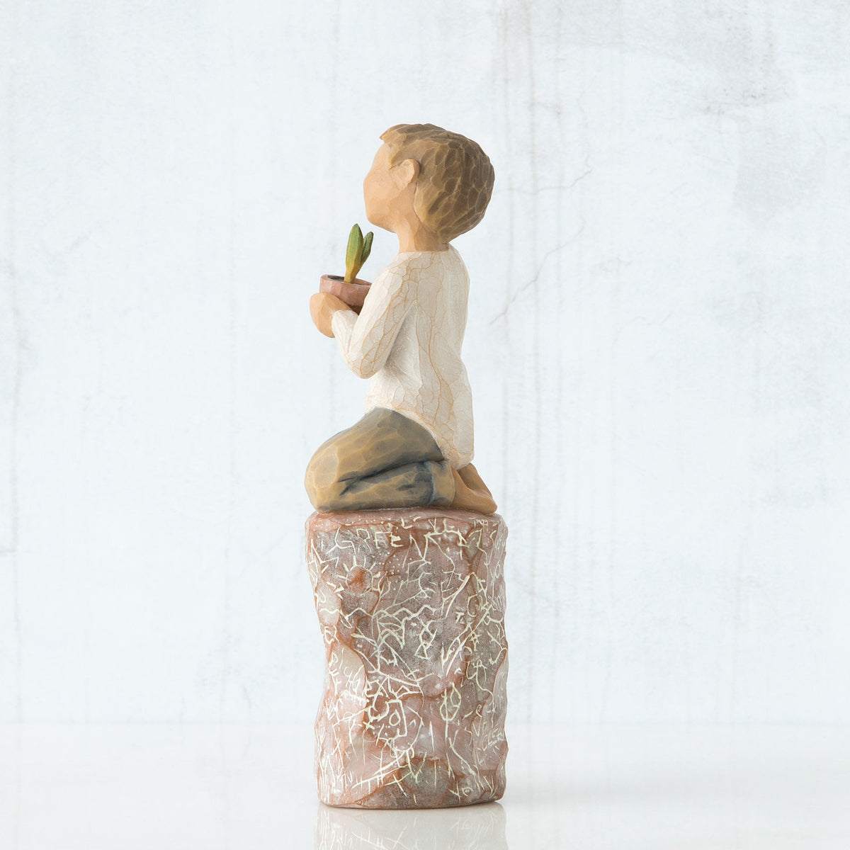 Right side view of boy with brown hair sitting on a stump while holding a small potted plant to his chest