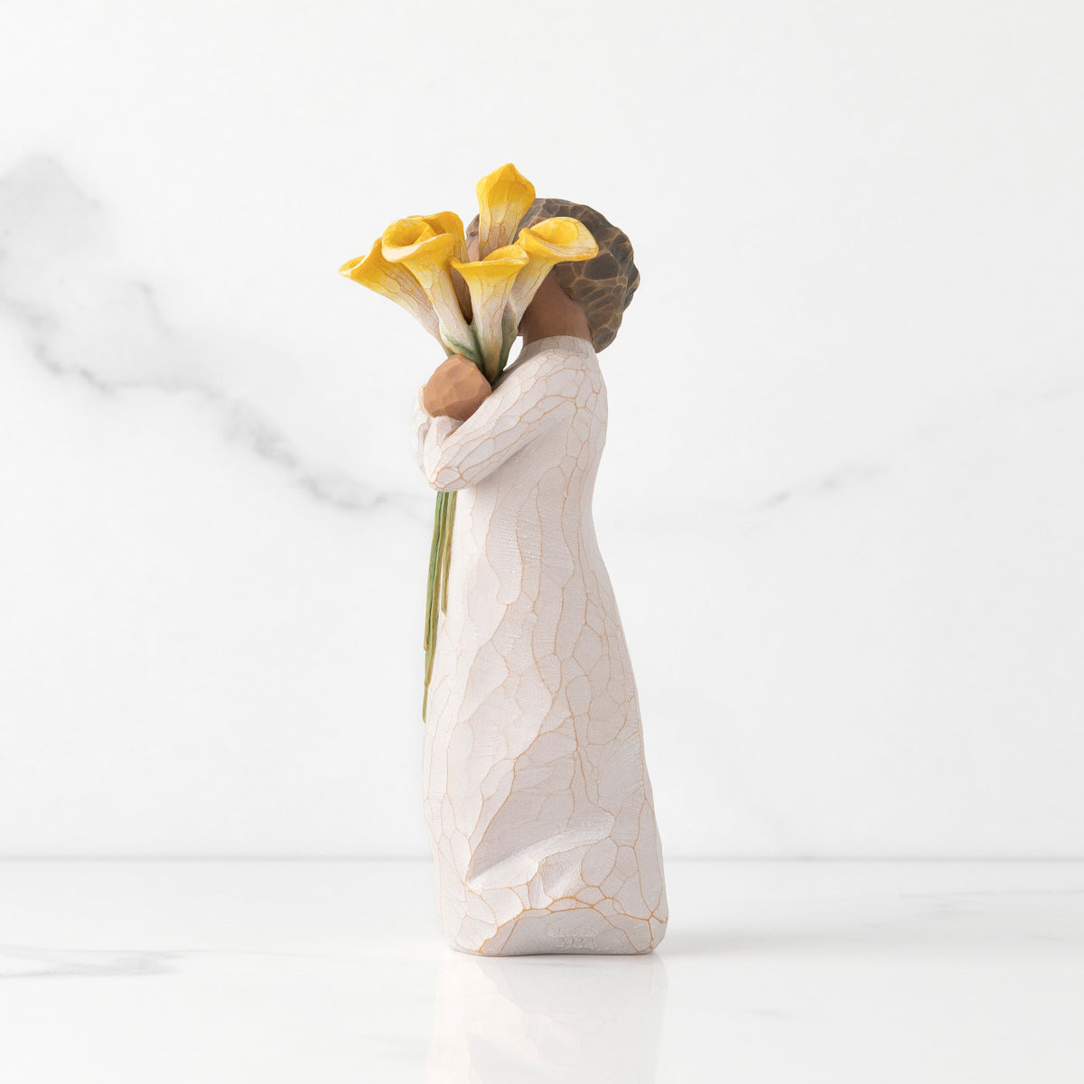 Left side of standing figure in cream dress with darker skin holding yellow calla lilies in her arms