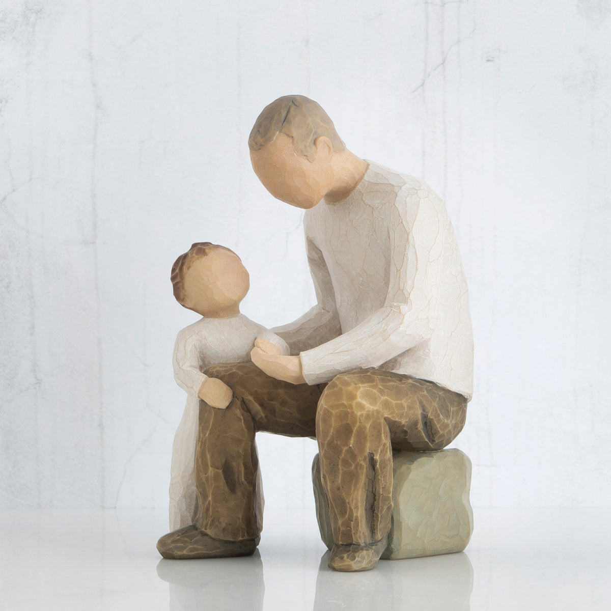 Man figurine in white shirt with brown pants sitting down with arm around little boy figurine