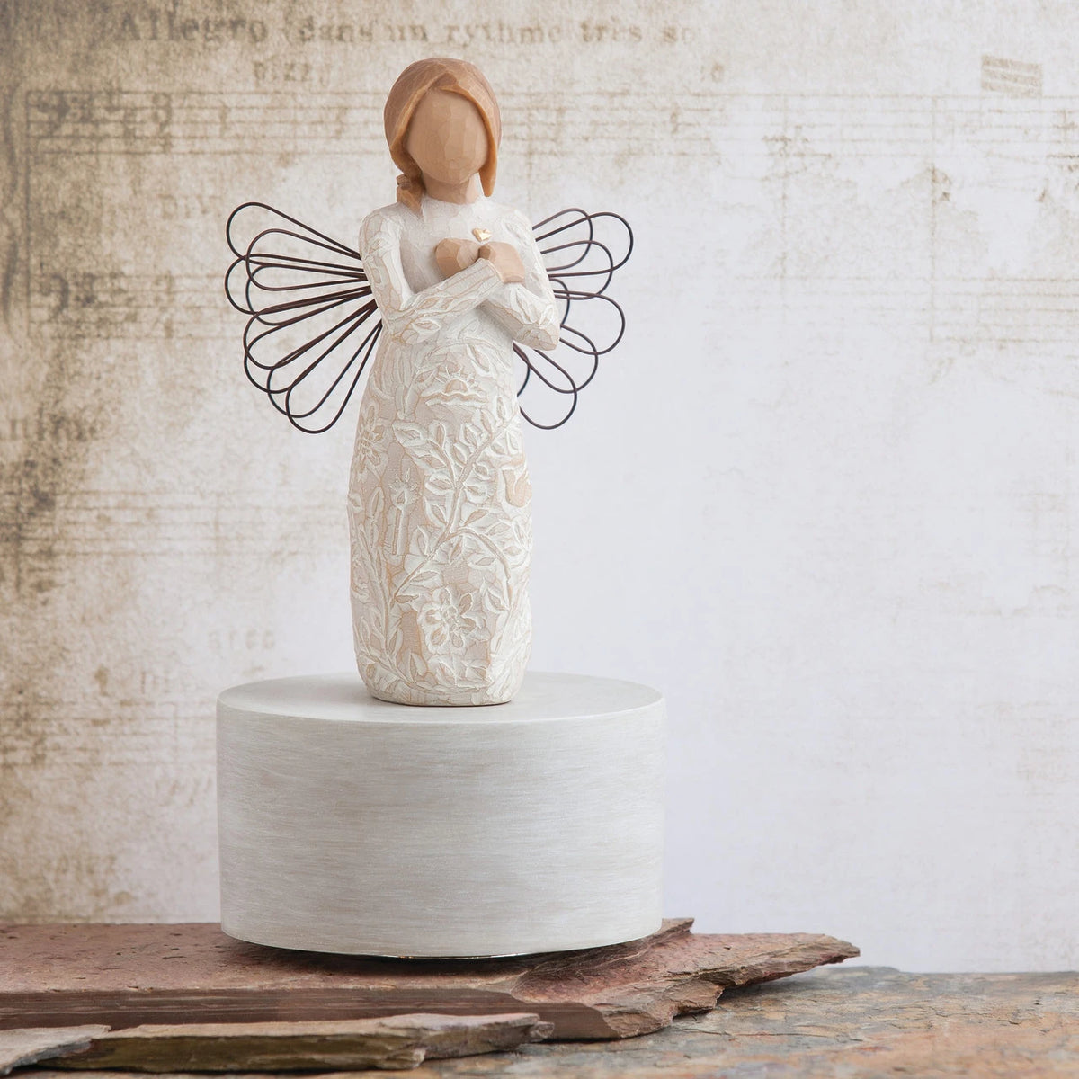 Willow Tree Remembrance Musical, 7.5”h hand-painted resin angel figure in cream dress carved with symbols of nature, wire wings, crossed hands under small gold-leaf heart, atop round, cream base containing musical element, in front of cream painting
