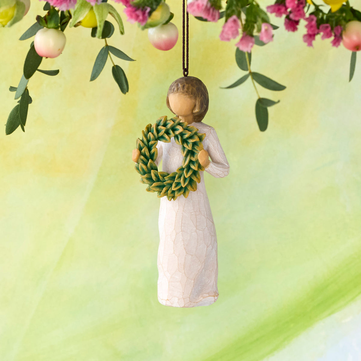 Willow Tree Magnolia Ornament, 4.25”h hand-painted resin figure in cream dress, holding green magnolia leaf wreath, brown elastic cord, hanging in front of pale green painting