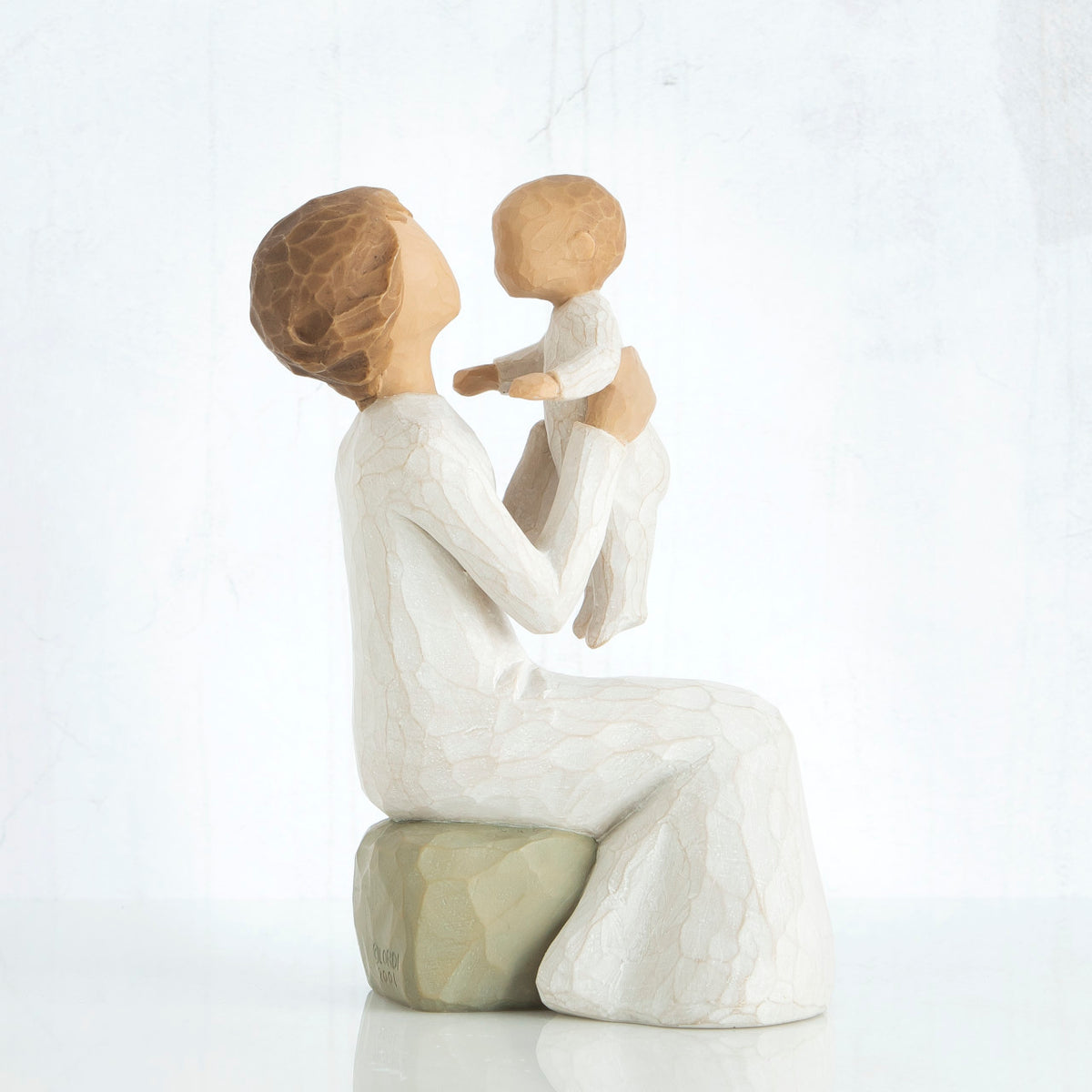Brunette mom figurine in white sitting on grey rock holding up baby figurine in her arms
