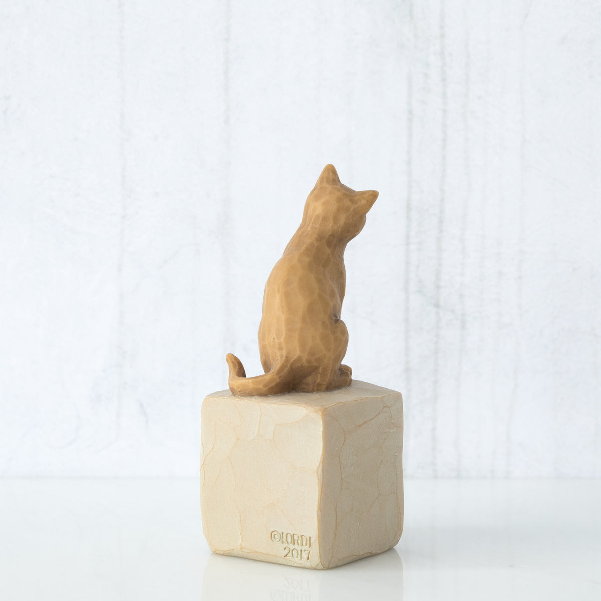 Figure of orange cat with tilted head perched atop a cream colored rectangular pedestal, facing back