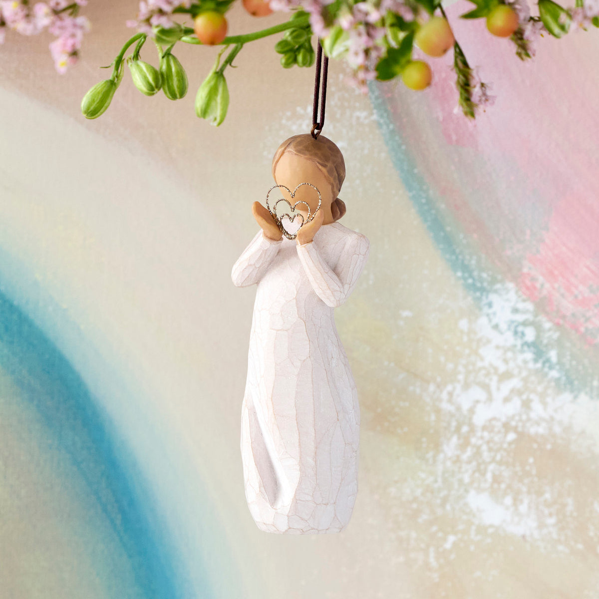 Willow Tree Lots of Love Ornament, 4.5”h hand-painted resin figure in cream dress, holding wire concentric heart with gold glitter, brown elastic cord, hanging in front of multi-pastel painting
