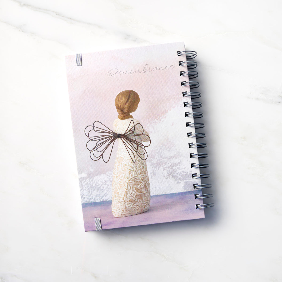 Back view: spiral-bound notebook featuring back of figure with wire wings in cream dress carved with natural symbols, hands crossed under gold heart on chest
