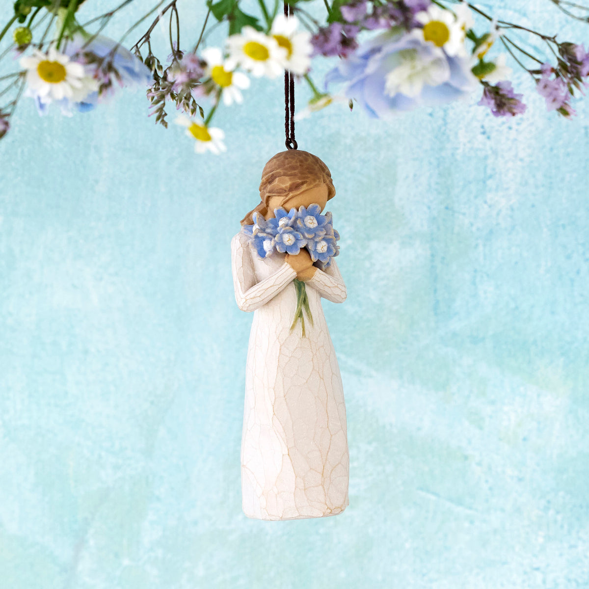 Willow Tree Forget-me-not Ornament, 4.25”h hand-painted resin figure in cream dress, holding bouquet blue flowers up to face, brown elastic cord, hanging in front of blue painting