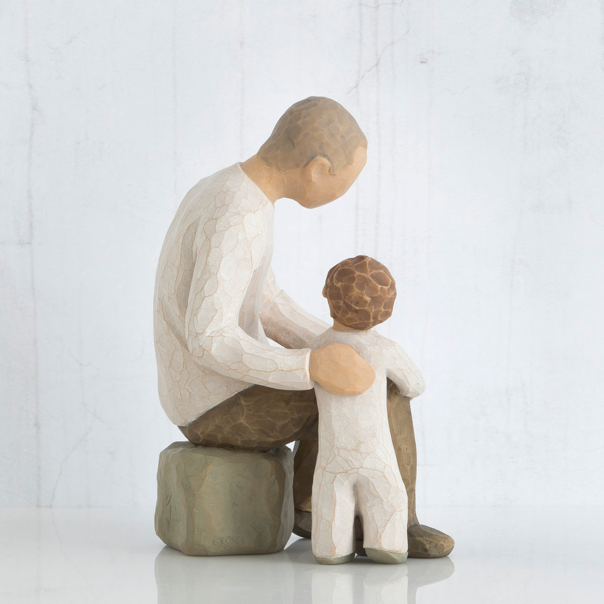 Father figurine in brown pants and white shirt sitting on rock with arm around little boy figurine in white