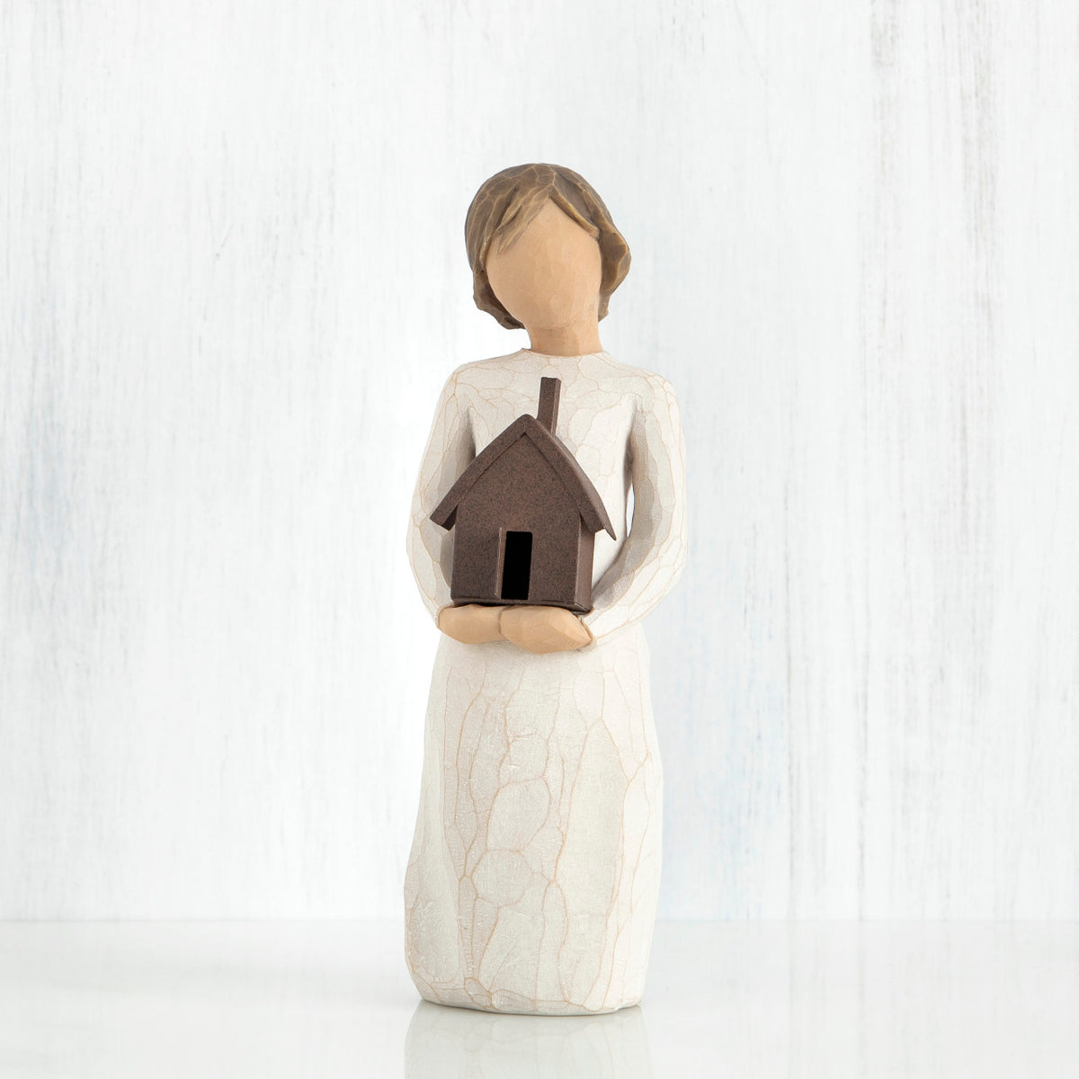 Faceless girl figurine facing forward wearing white dress, holding brown house figurine in her arms