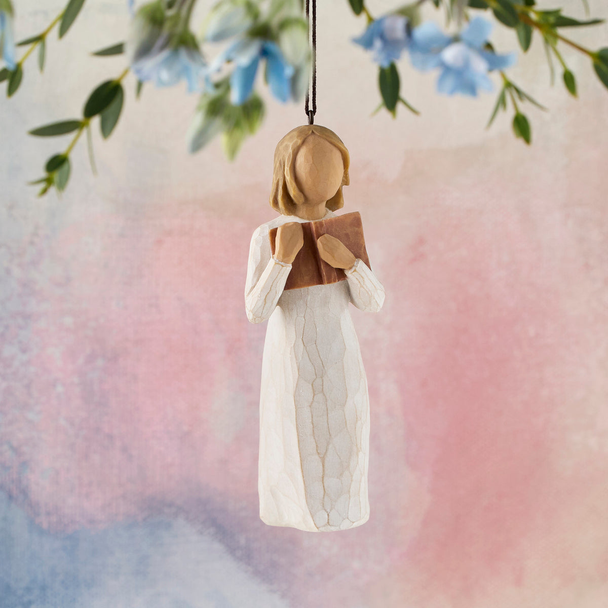 Willow Tree Love of Learning Ornament, 4”h hand-painted resin figure in cream dress holding open book to her chest, brown elastic cord, hanging in front of pink painting