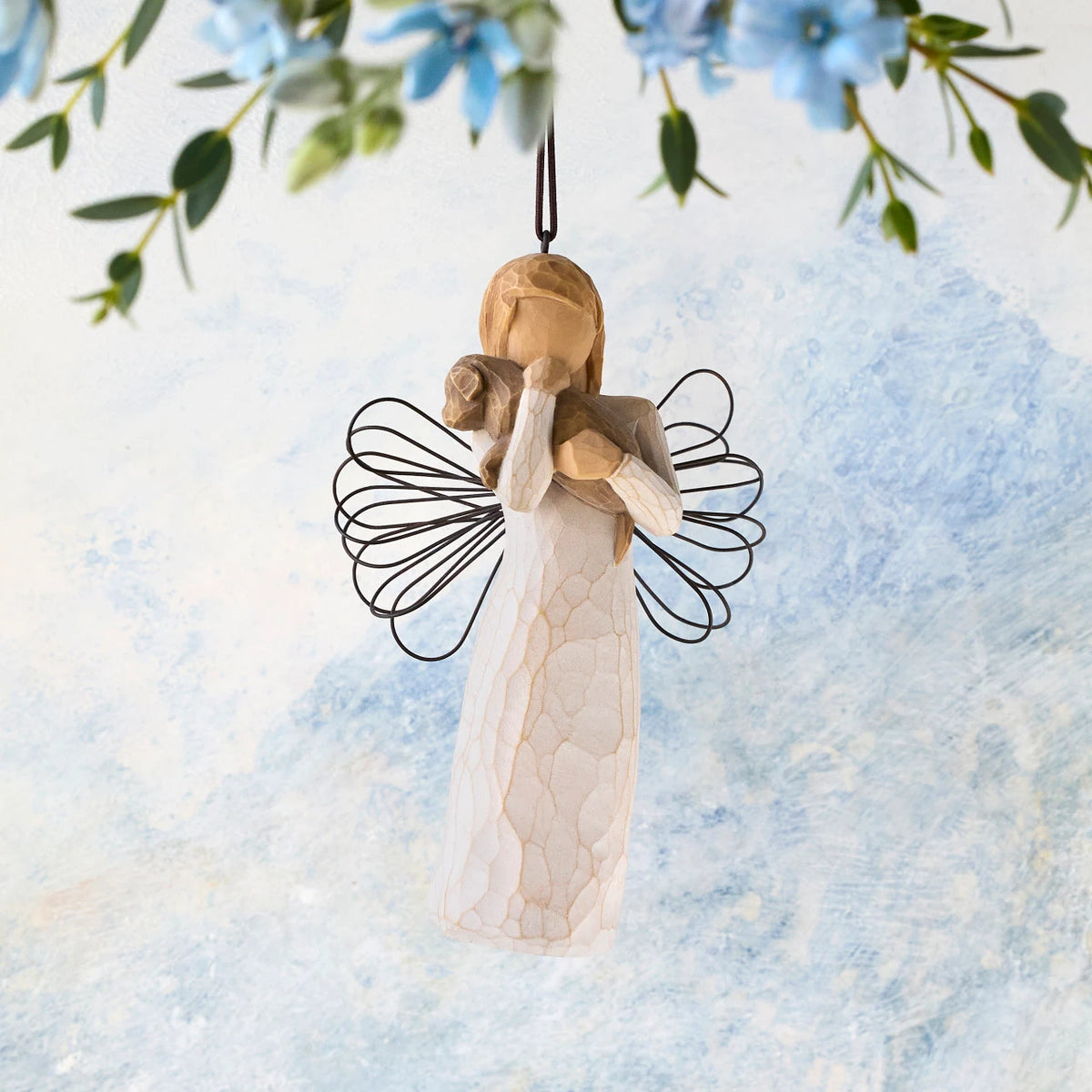 Willow Tree Angel of Friendship Ornament, 4.5"h hand-painted resin figure in cream dress with wire wings holding brown dog, brown elastic cord, hanging in front of sky blue painting
