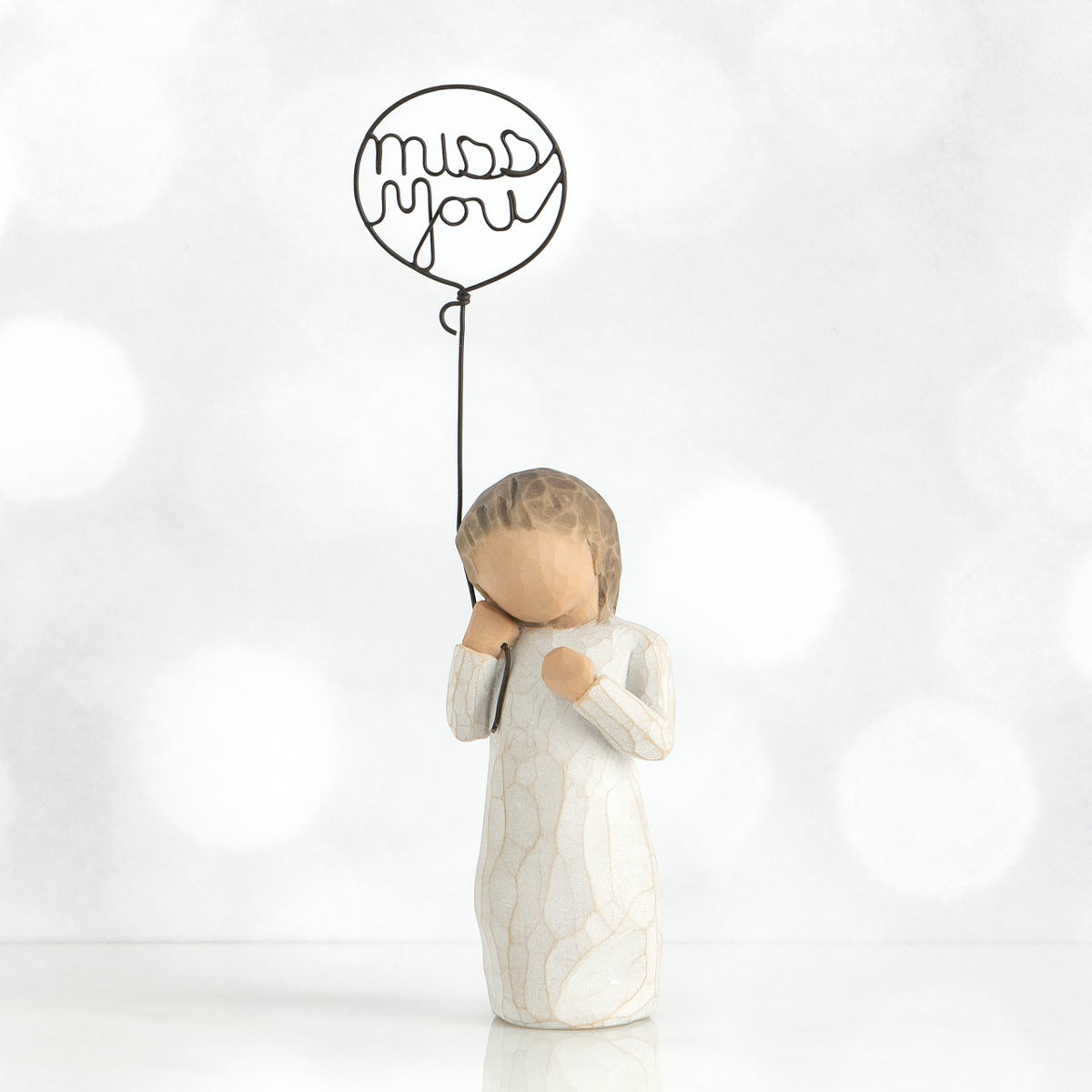 Small faceless girl figurine in white dress holding 'miss you' black wire balloon