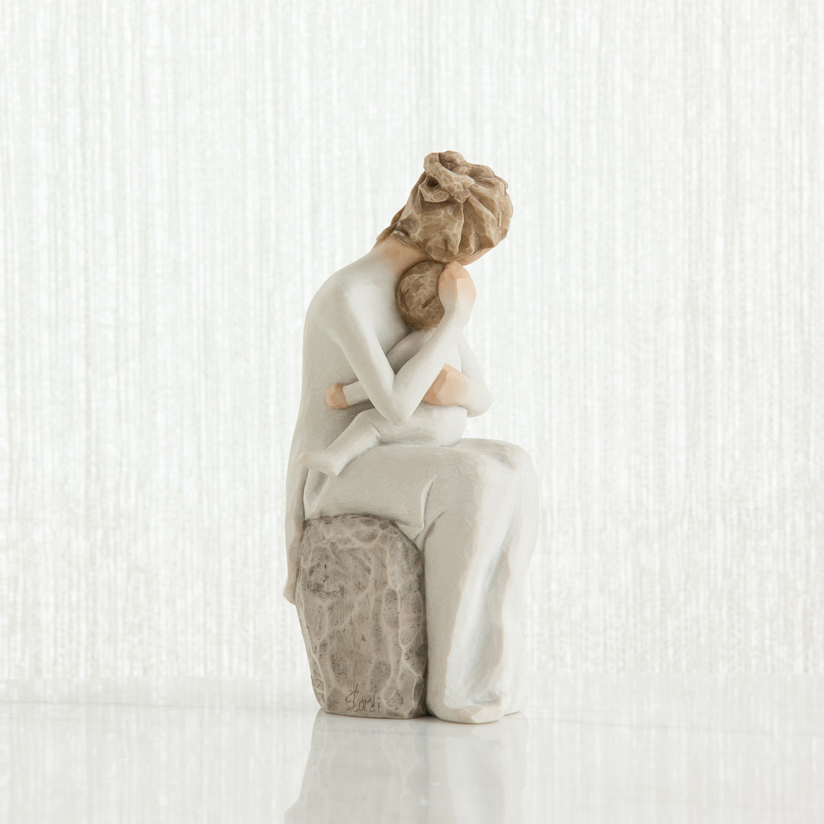 Woman figurine sitting on grey rock dressed in white holding small baby facing away from the front - white background