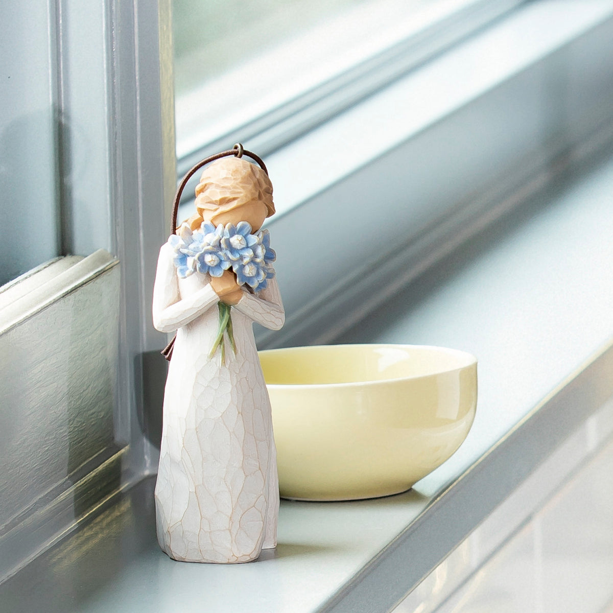 Willow Tree Forget-me-not Ornament, 4.25”h hand-painted resin figure in cream dress, holding bouquet blue flowers up to face, brown elastic cord, on bright windowsil