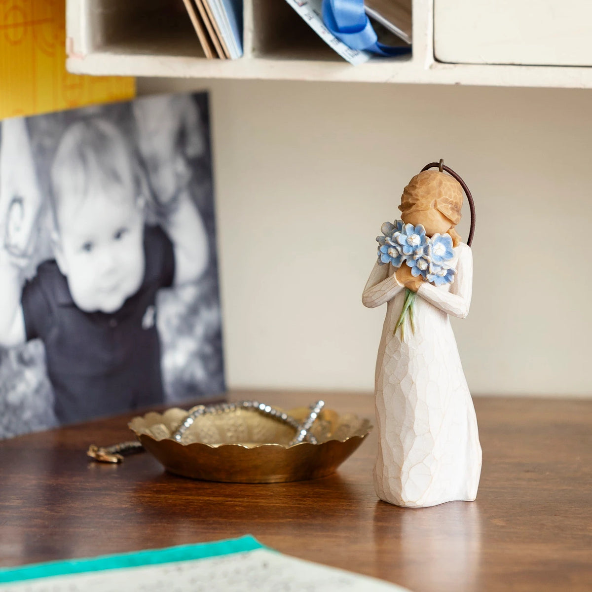Willow Tree Forget-me-not Ornament, 4.25”h hand-painted resin figure in cream dress, holding bouquet blue flowers up to face, brown elastic cord, on desk with photos