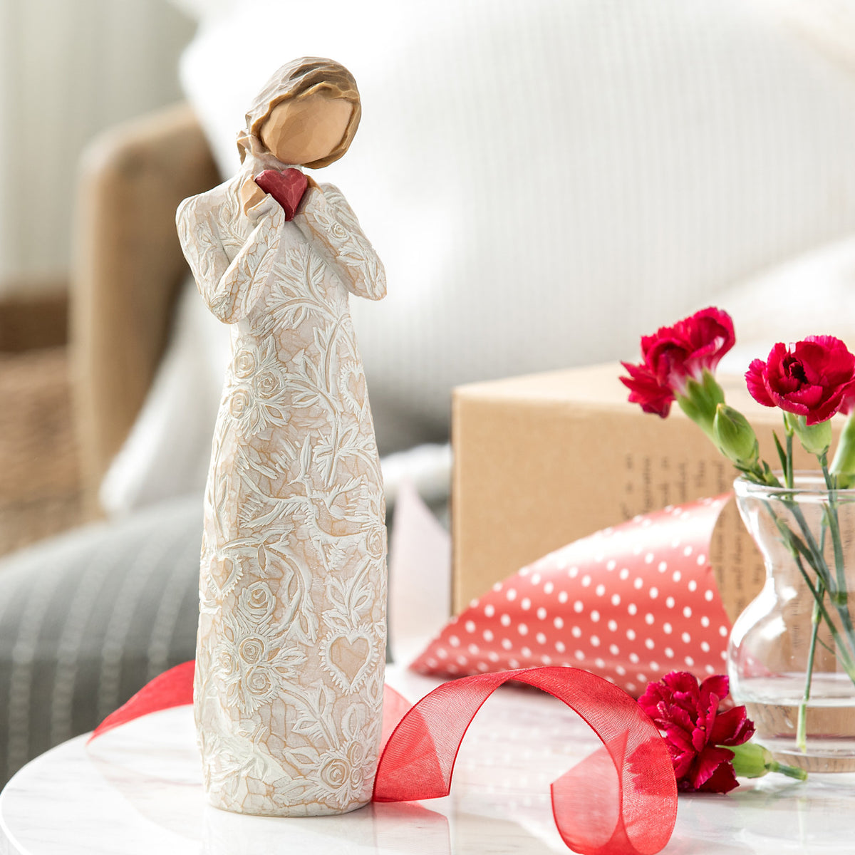 Willow Tree Je t'aime (I Love You), 8.5"h hand-painted standing resin figure holding red heart in cream dress with ruffled neck and carved with hearts, on table with red flowers