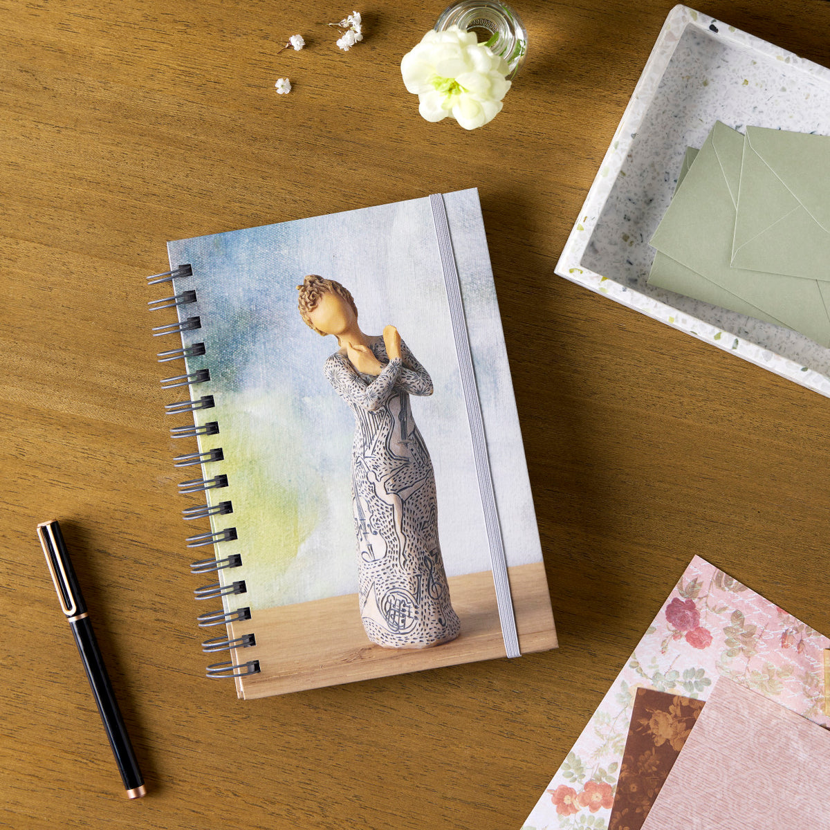 Willow Tree Music Speaks Notebook, 6.5"w x 8.5"h, hard-cover, spiral-bound, 240 lined pages, front cover features Music Speaks figure on background of blue-green painting, closed on desk