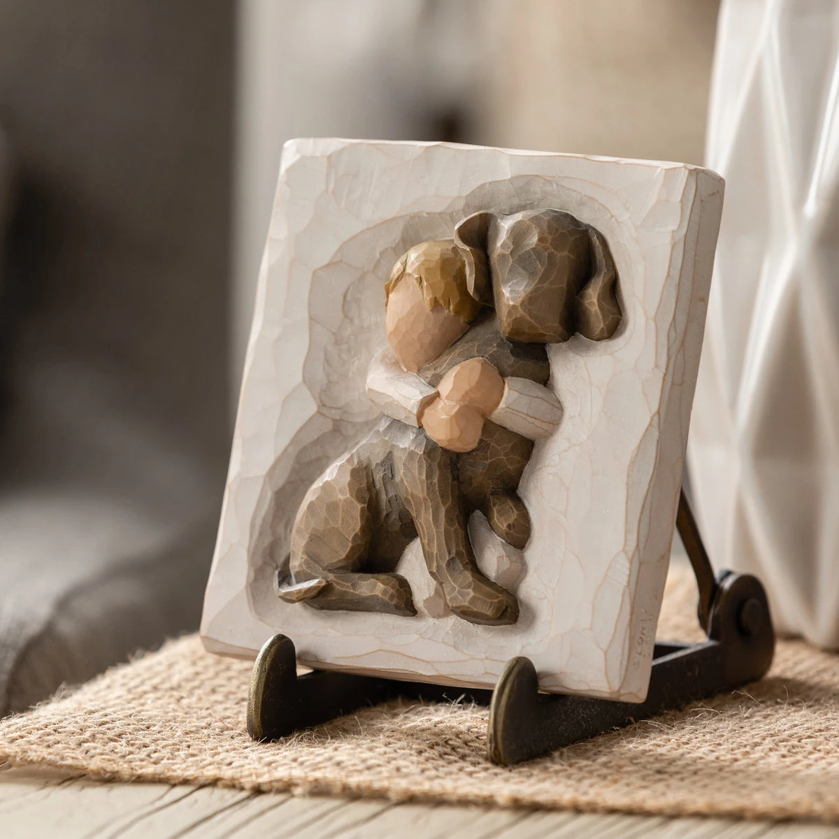 Willow Tree Hug Plaque, 4”sq x .5”d hand-painted resin bas-relief carving of small boy in cream shirt, hugging large dark dog. Plaque displayed on Plaque Stand on table