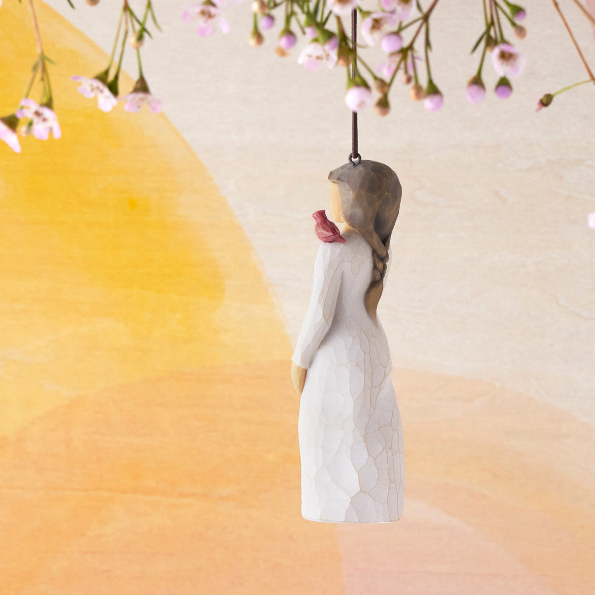 Willow Tree Messenger Ornament, side view of 4”h hand-painted resin figure in cream dress with red cardinal on shoulder, brown elastic cord, hanging in front of orange painting