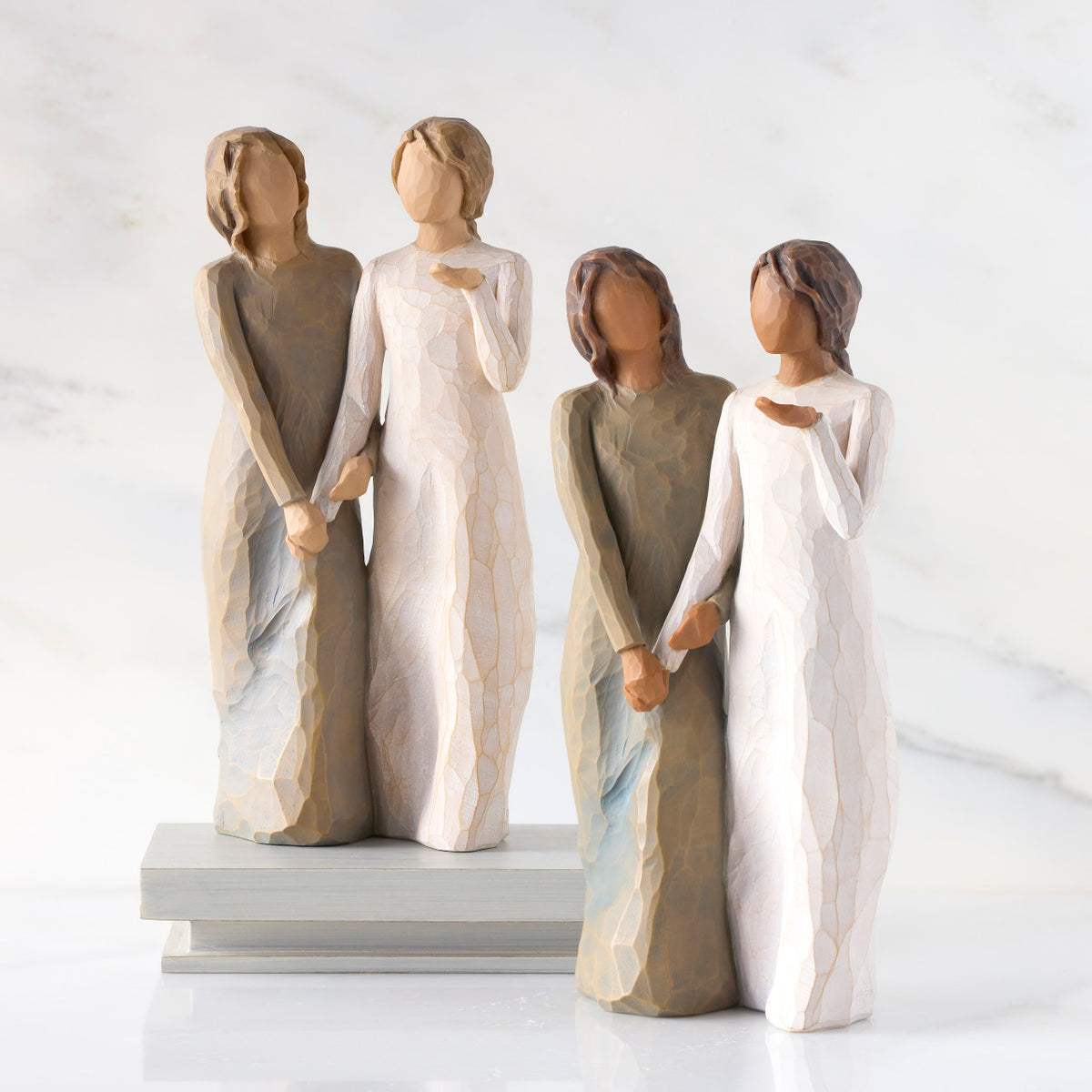 Lighter &amp; Darker skin versions of two standing female figures, one in cream dress, one in blue-gray dress, holding hands and turned as if speaking to each other