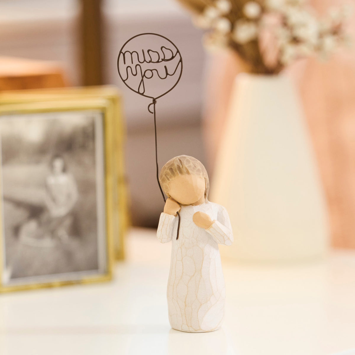 Willow Tree Miss You, 5”h hand-painted standing resin figure in cream dress holding wire ballon with words 'miss you' written in wire, on table with photo frames